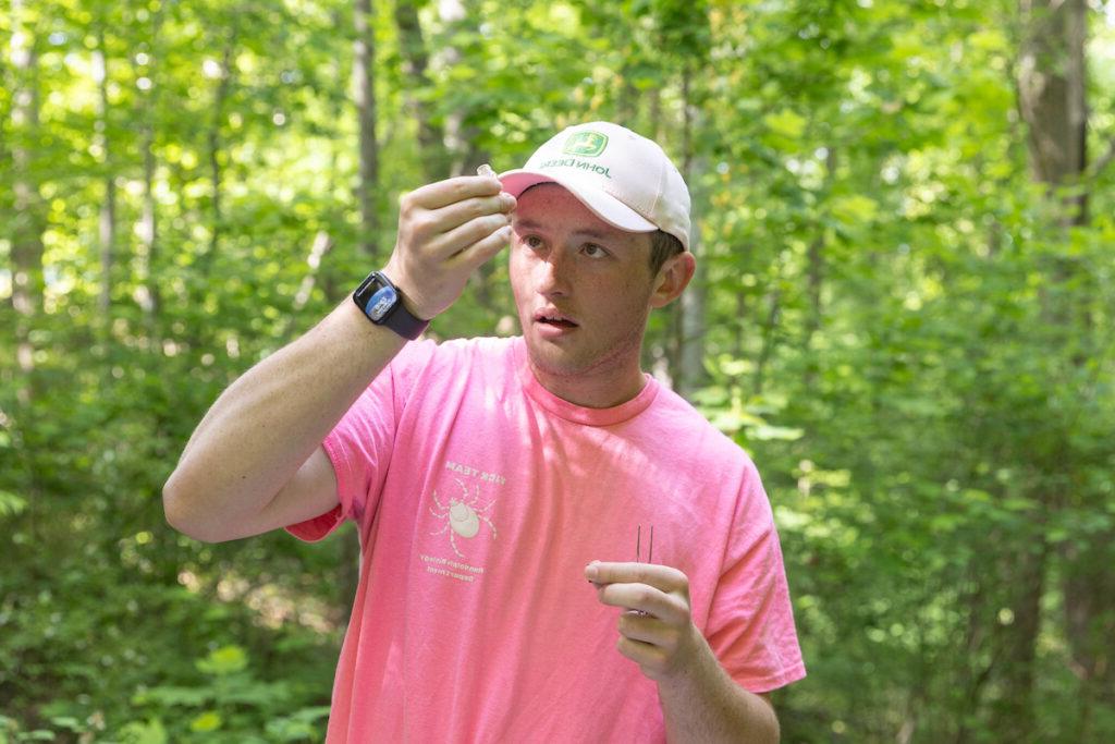 Ethan Caldwell holds up a tick while standing in a wooded area.
